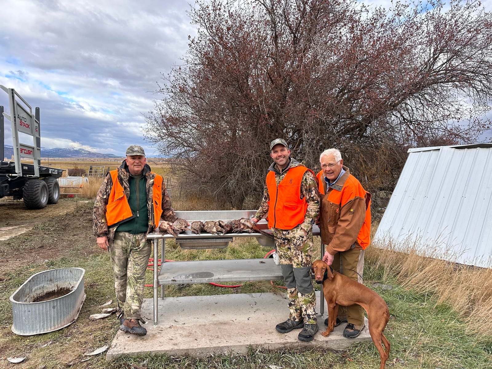 Three hunters and a dog pose with harvested ring-necked pheasants on a table outdoors.