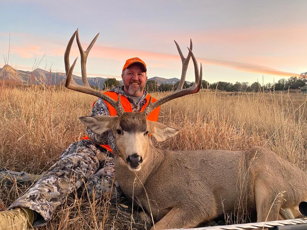 A hunter poses with a harvested mule deer in a field at sunset.