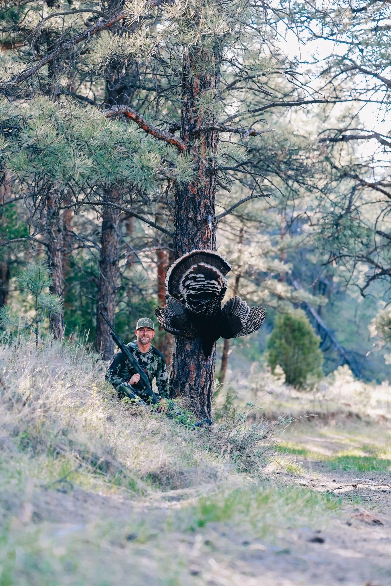 A hunter poses with a harvested wild turkey in a forest.
