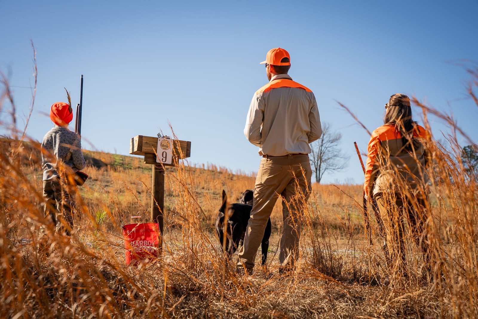 Three hunters and a dog stand in a field of tall, dry grass under a clear sky.