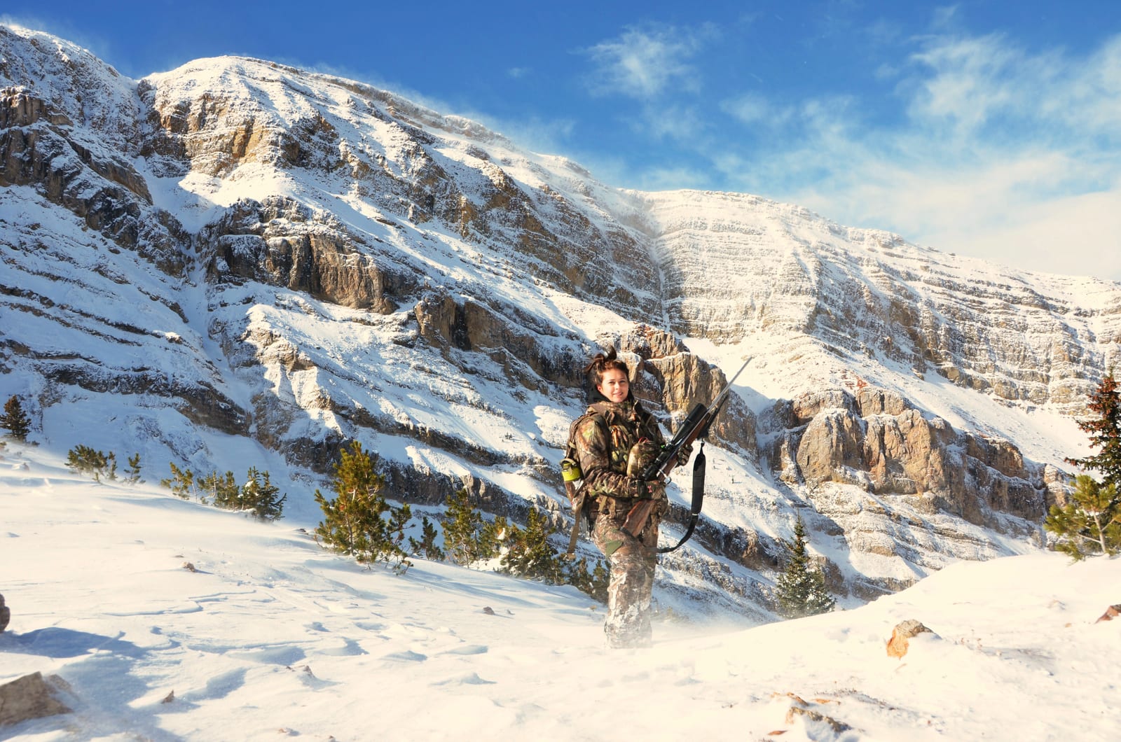 A hunter stands with a rifle in a snowy mountain landscape.
