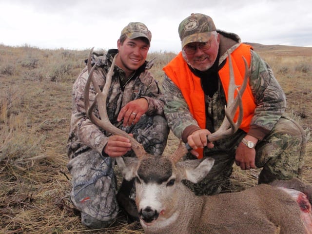 Two hunters pose with a harvested mule deer in a field.
