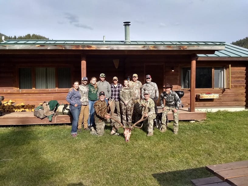 A group of hunters and guides pose with a harvested elk in front of a wooden lodge.