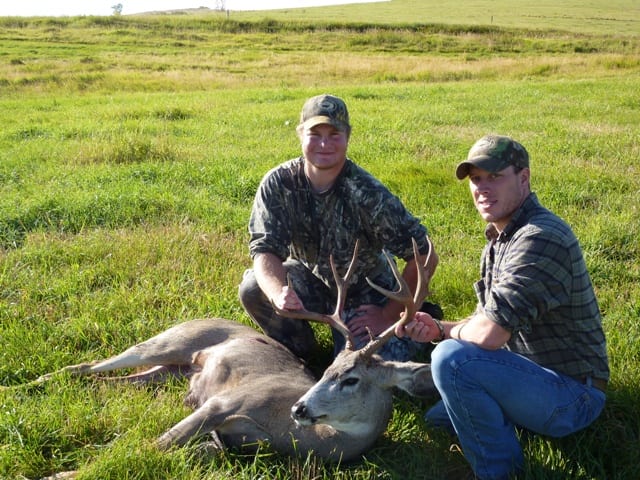 Two hunters pose with a harvested whitetail deer in a grassy field.