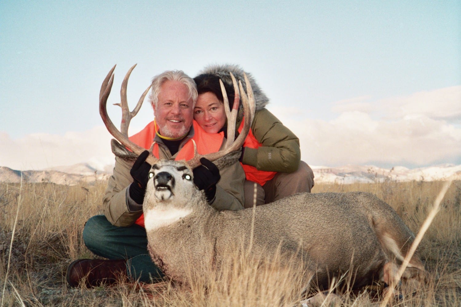 Two hunters pose with a harvested mule deer in a grassy field.