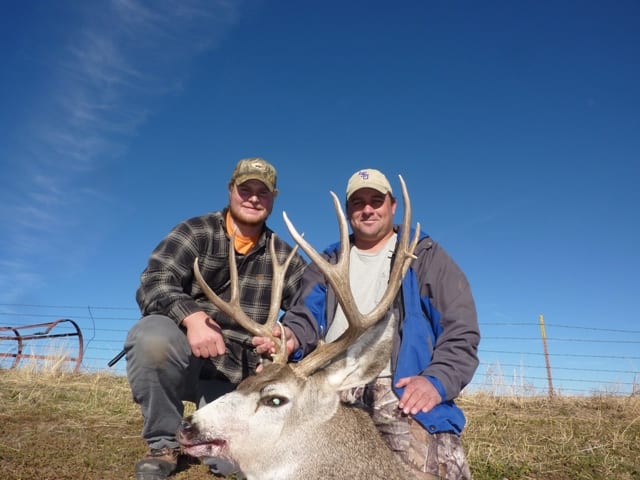 Two hunters pose with a harvested mule deer under a clear blue sky.