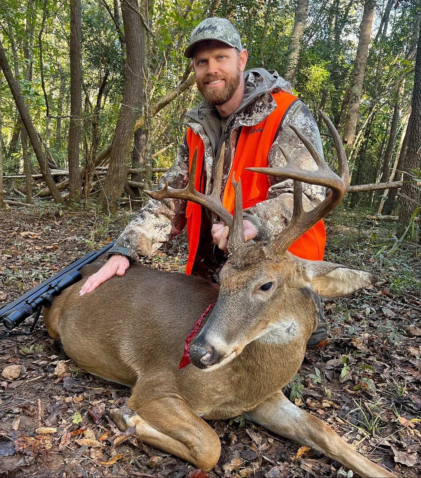 A man kneeling on the forest floor, smiling and holding the antlers of a recently hunted deer with large antlers, in a wooded area surrounded by green trees.