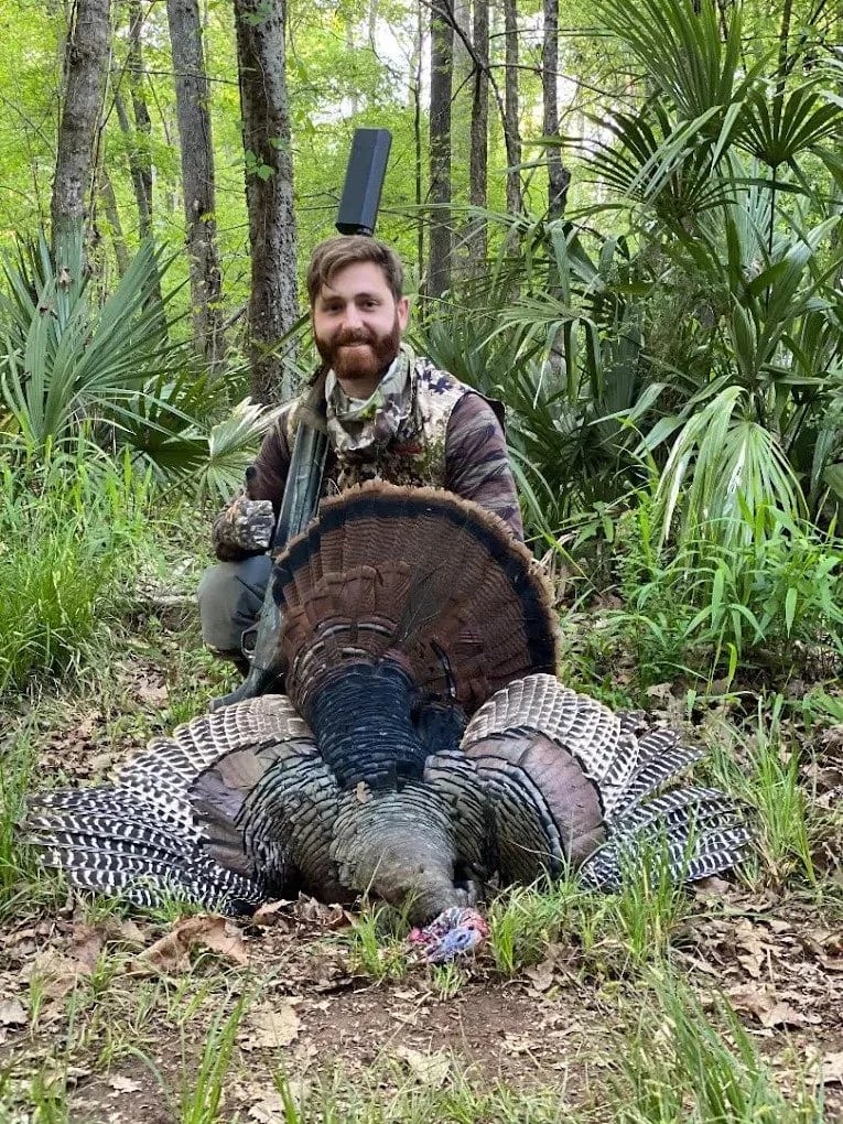 A man with a beard in camouflage clothes kneeling in a forest while holding a hunting rifle. In front of him lies a large fallen turkey with its feathers spread out on the ground.
