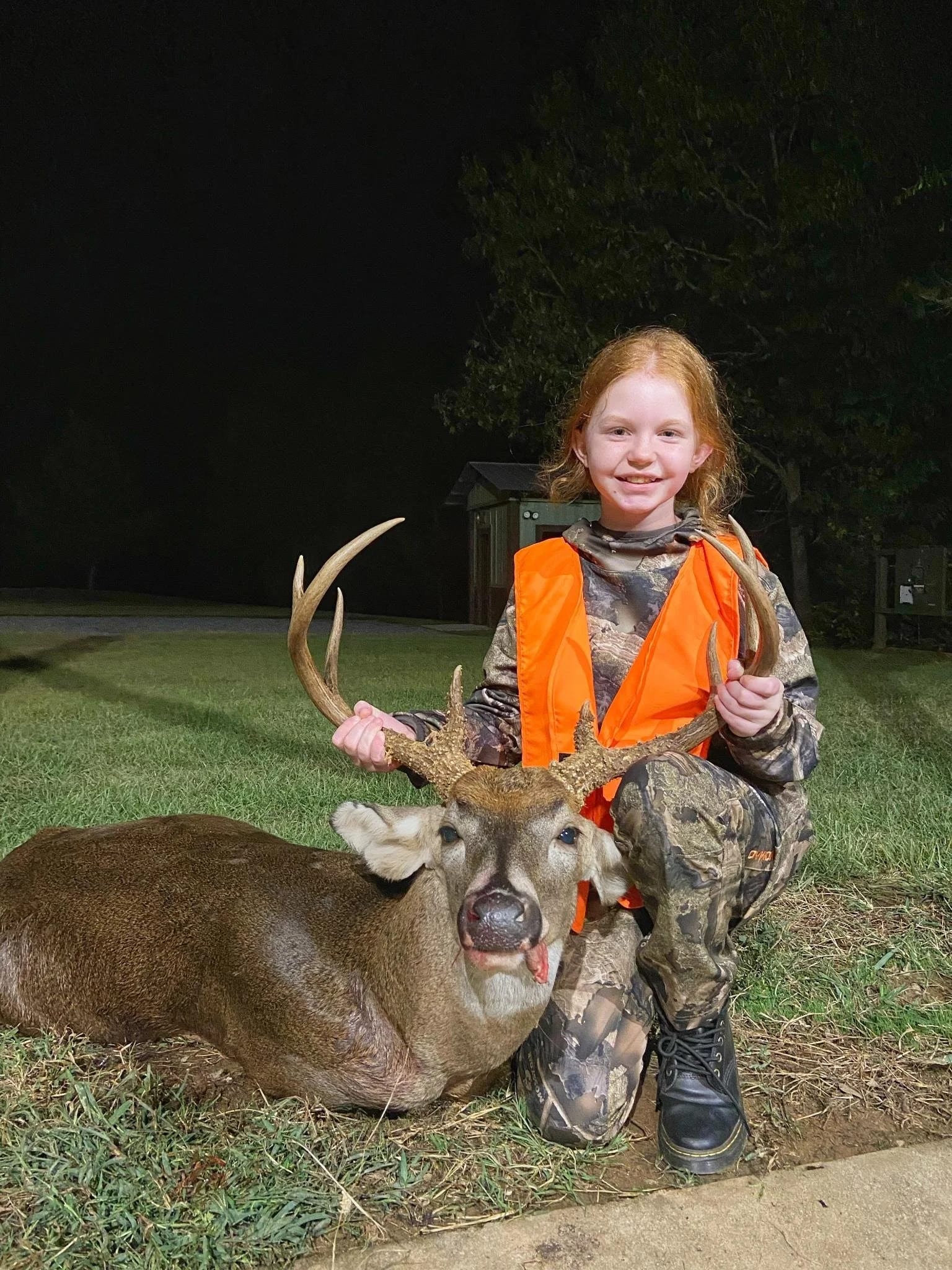A young girl with red hair in camouflage clothing and orange safety vest kneeling next to a large deer with antlers, holding its antlers, at night outdoors with grass and trees in the background.