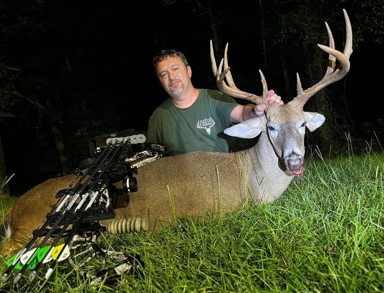 A man with a goatee and wearing a green T-shirt is posing with a large deer he has hunted. The deer is lying on the grass, and the man is holding its antlers. The setting is outdoors at night, with dark trees in the background.