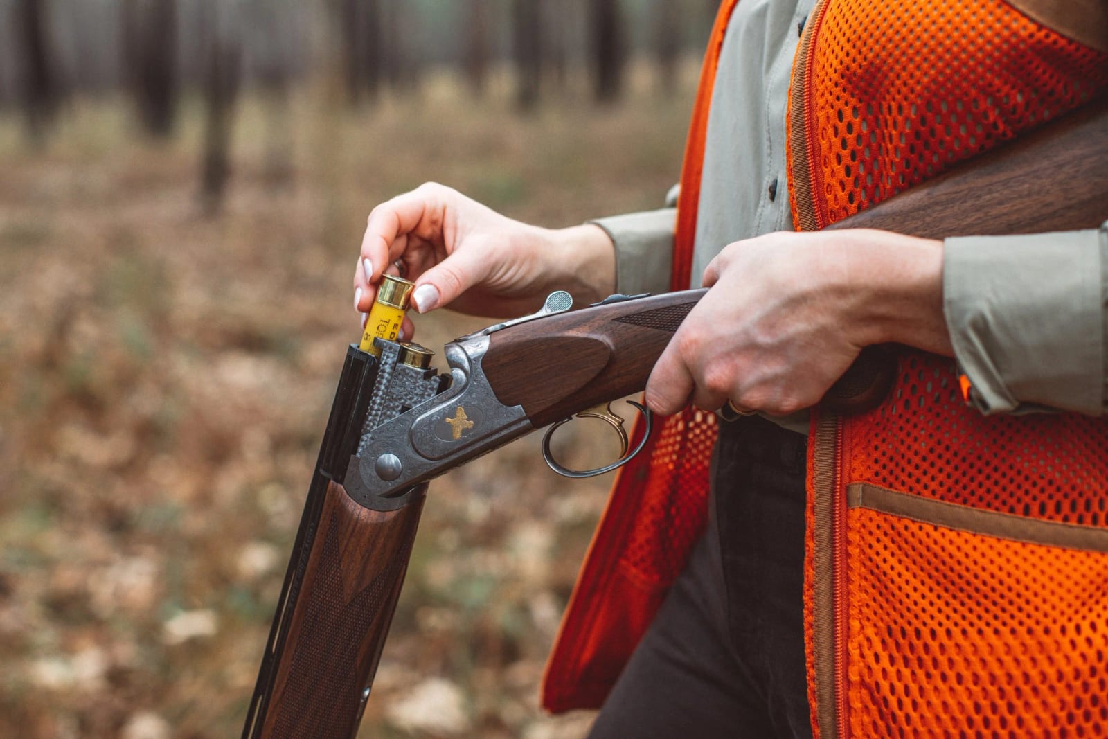 A person in an orange hunting vest loads a shotgun with a yellow shell.