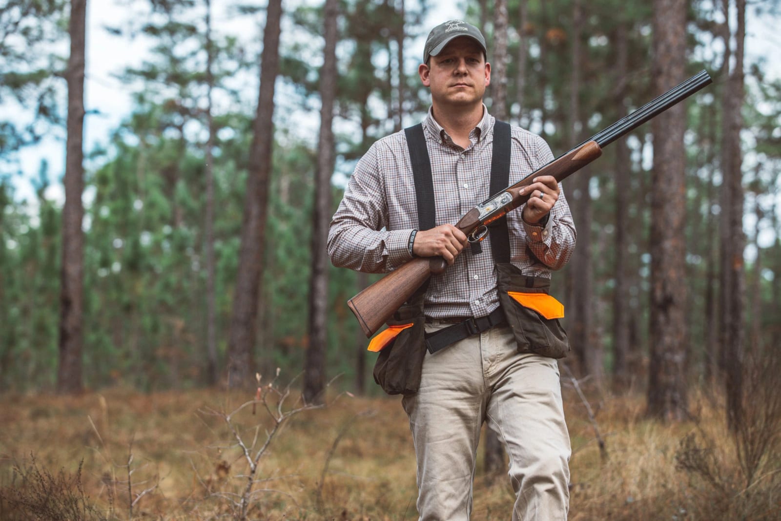 A man in hunting attire holds a shotgun in a pine forest.