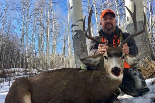 A hunter poses with a harvested mule deer in a snowy, wooded area.