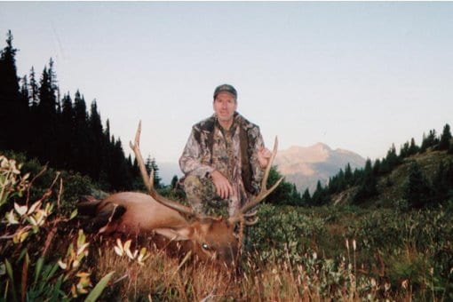 A hunter in camouflage kneels next to a harvested elk in a mountainous, forested landscape.