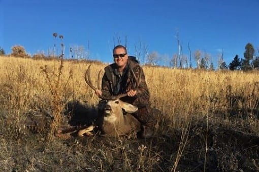 A hunter in camouflage poses with a harvested mule deer in a grassy field under a clear blue sky.