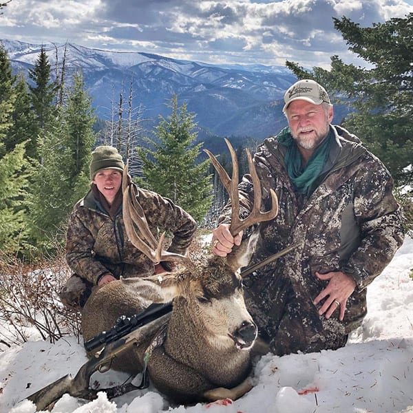Two hunters pose with a harvested mule deer in a snowy, mountainous landscape.