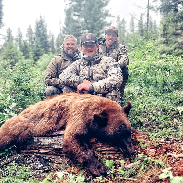 Three hunters pose with a harvested black bear in a forest.