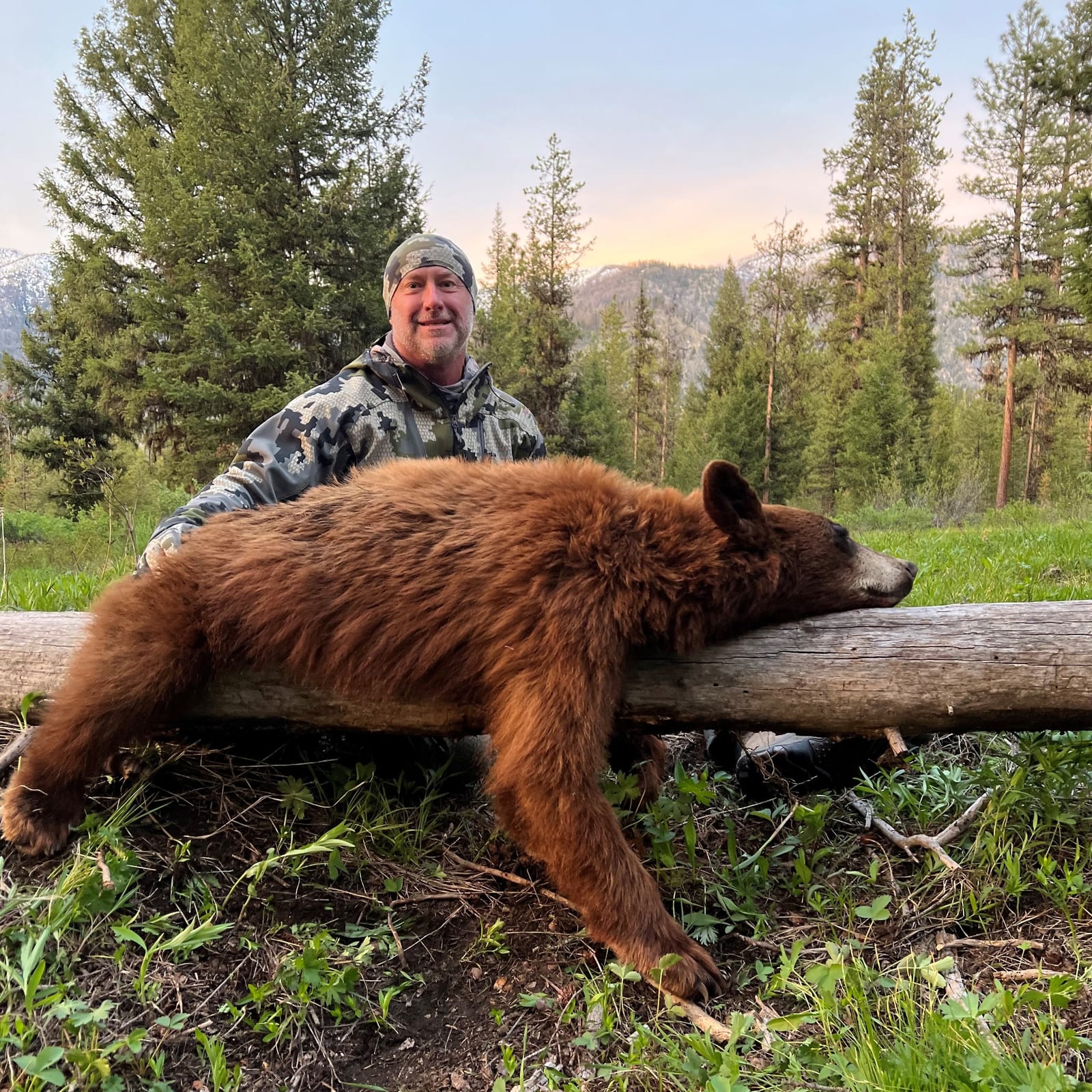 A hunter poses with a harvested black bear on a log in a forest.