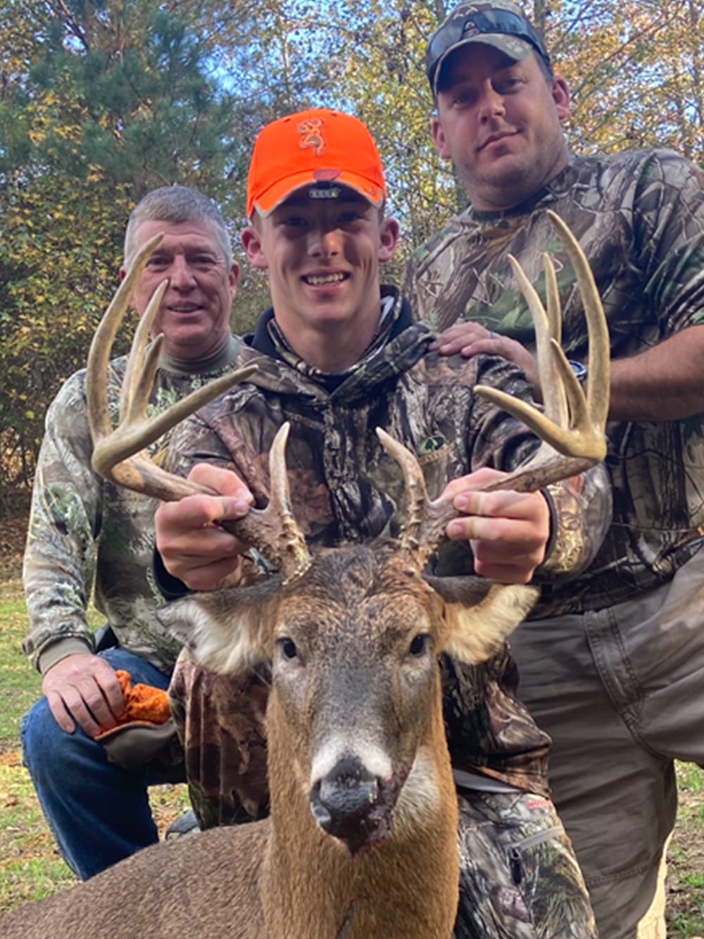 Three men in camouflage pose with a harvested whitetail deer.