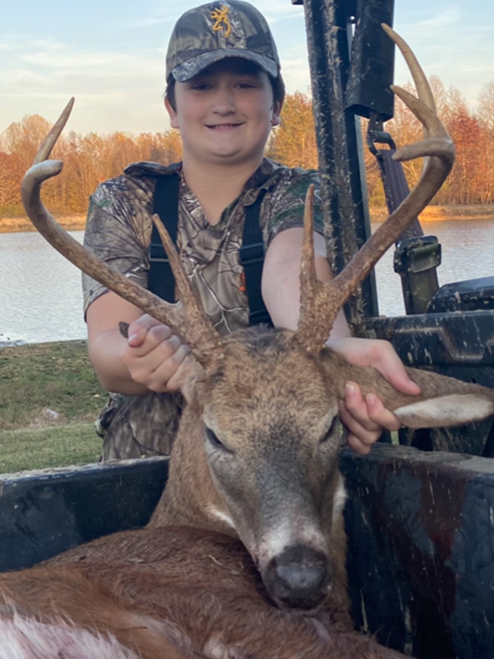 A young hunter poses with a harvested whitetail deer in the back of a vehicle, with a body of water and trees in the background.