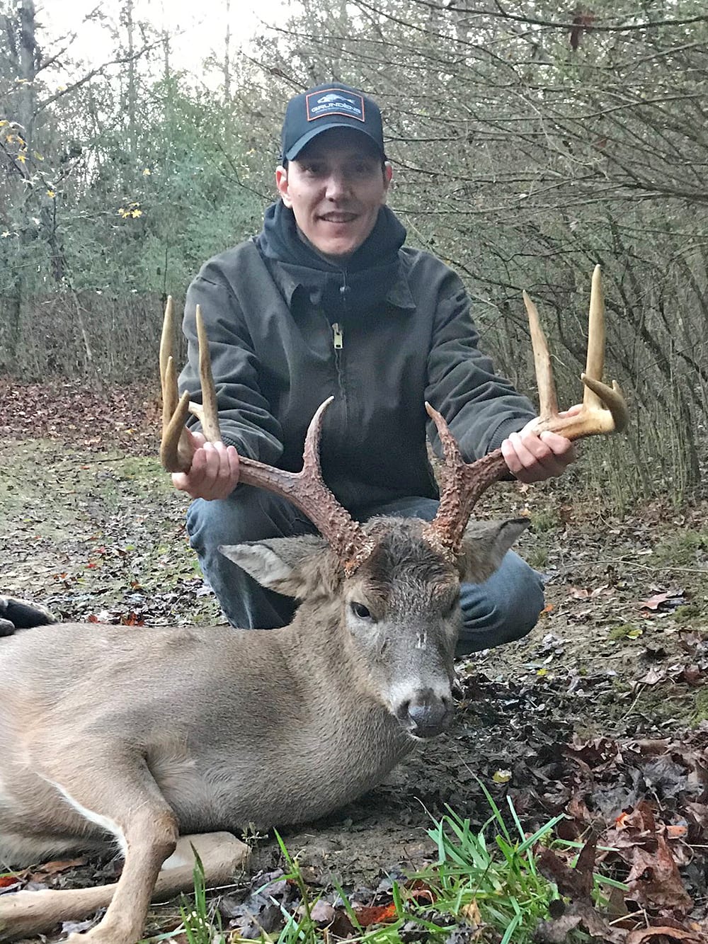 A hunter kneels behind a harvested whitetail deer, holding its antlers, in a wooded area.