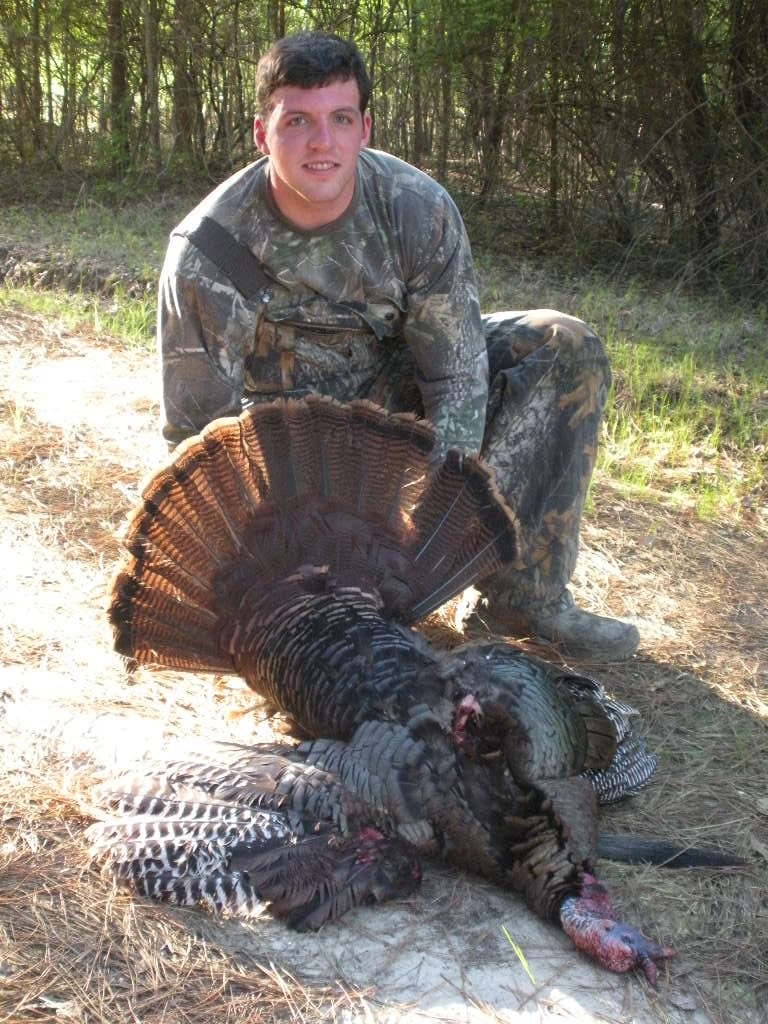 A hunter in camouflage kneels next to a harvested wild turkey with its tail fanned out.