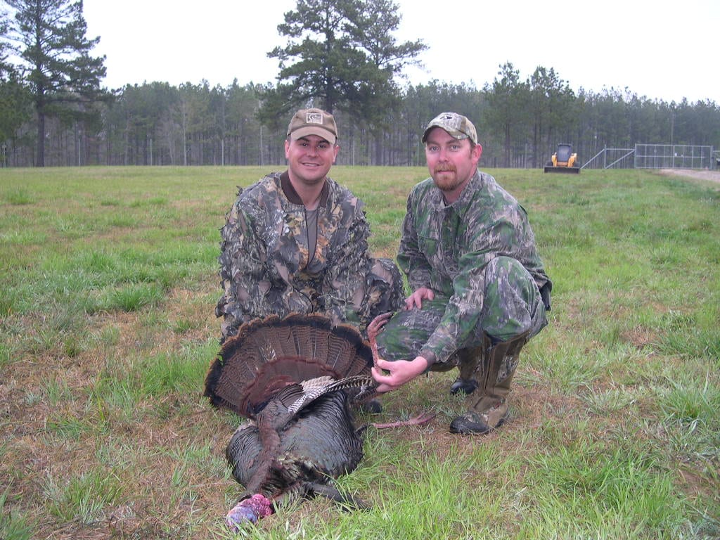 Two hunters in camouflage pose with a harvested wild turkey in a grassy field.