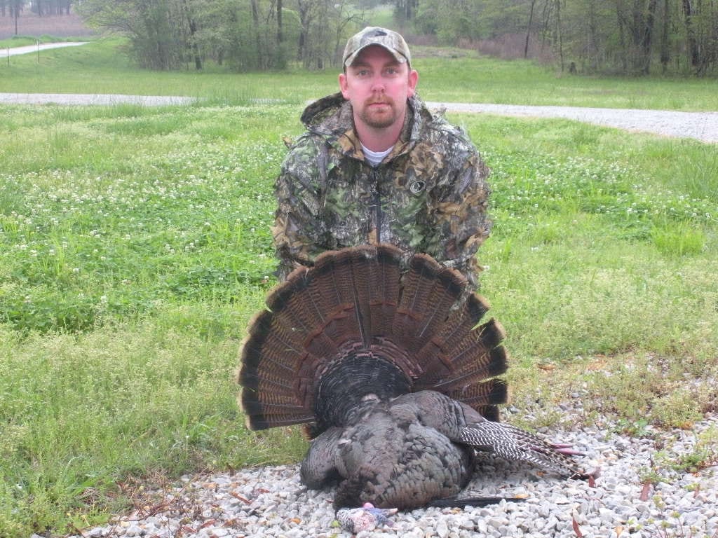 A hunter in camouflage poses with a harvested wild turkey in a grassy field.