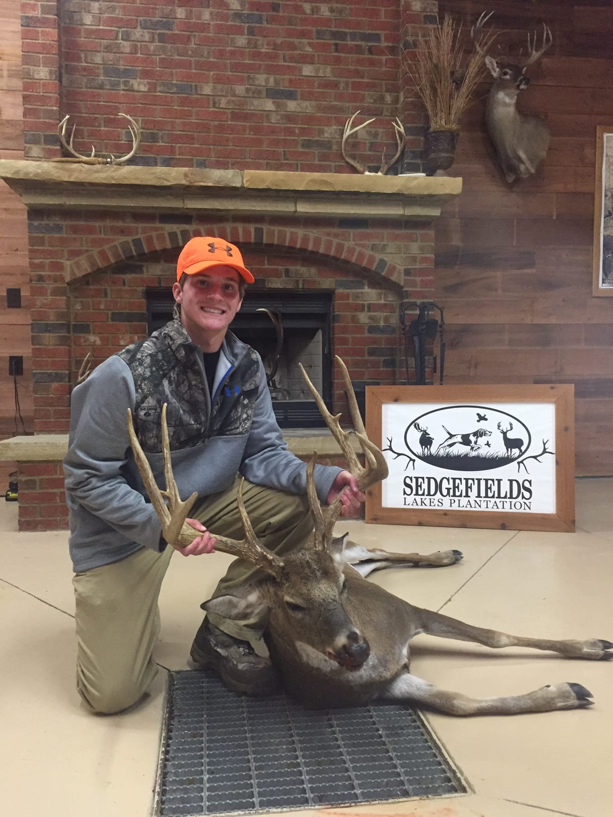 A hunter kneels next to a harvested whitetail deer indoors, with a fireplace and a 'Sedgefields Lakes Plantation' sign in the background.