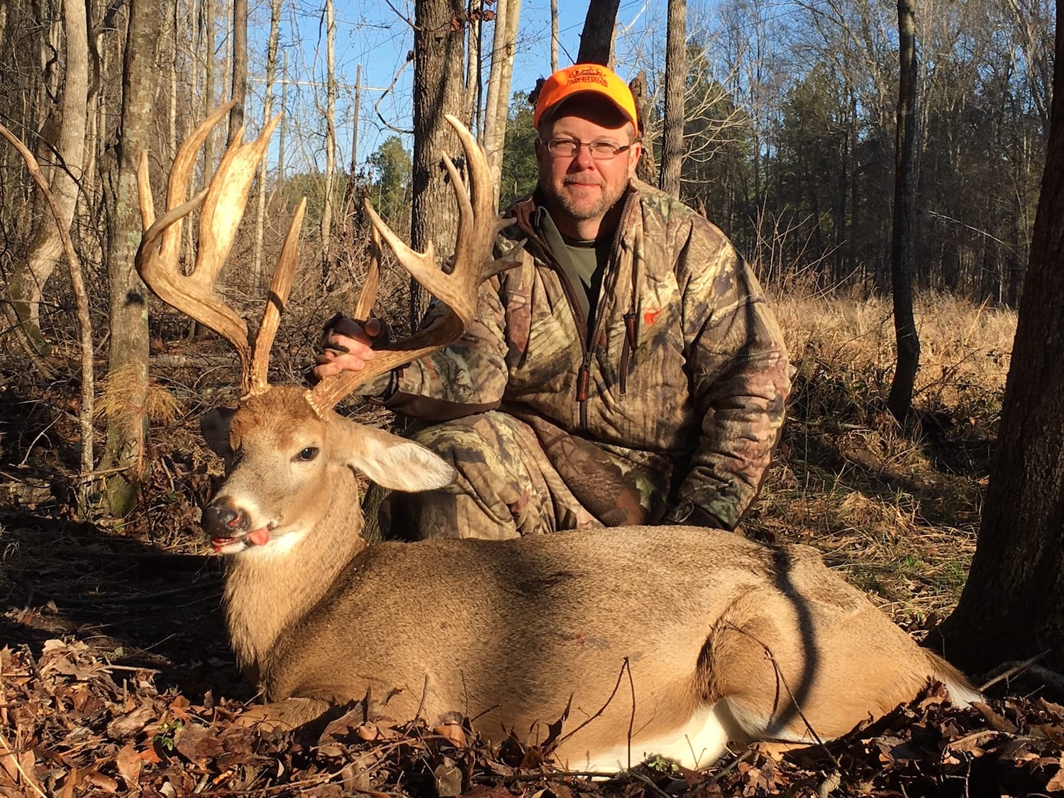 A hunter poses with a harvested whitetail deer in a wooded area.