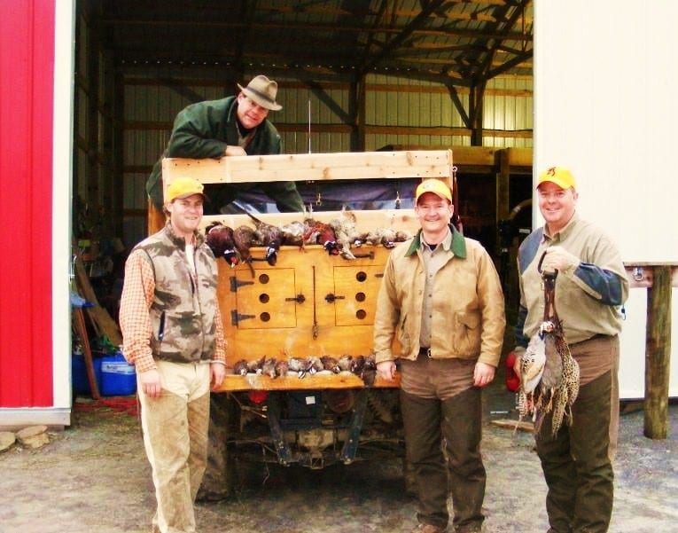 Three men pose with harvested ring-necked pheasants in front of a wooden game cart.