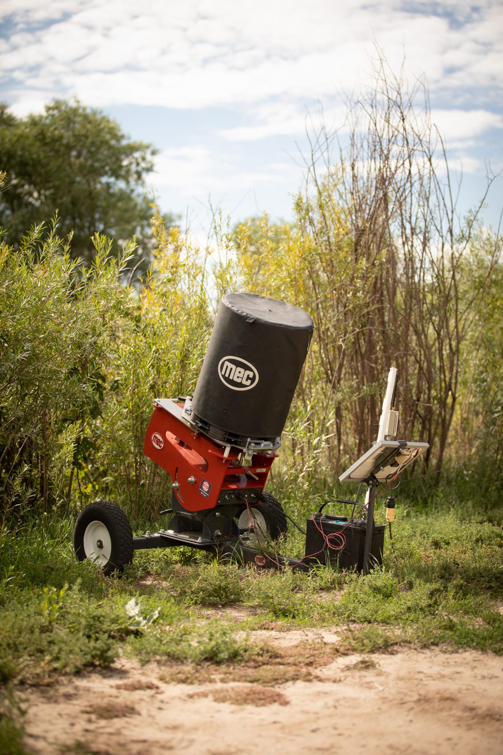 A MEC clay target thrower with a solar panel setup is positioned in a grassy area with trees under a cloudy sky.