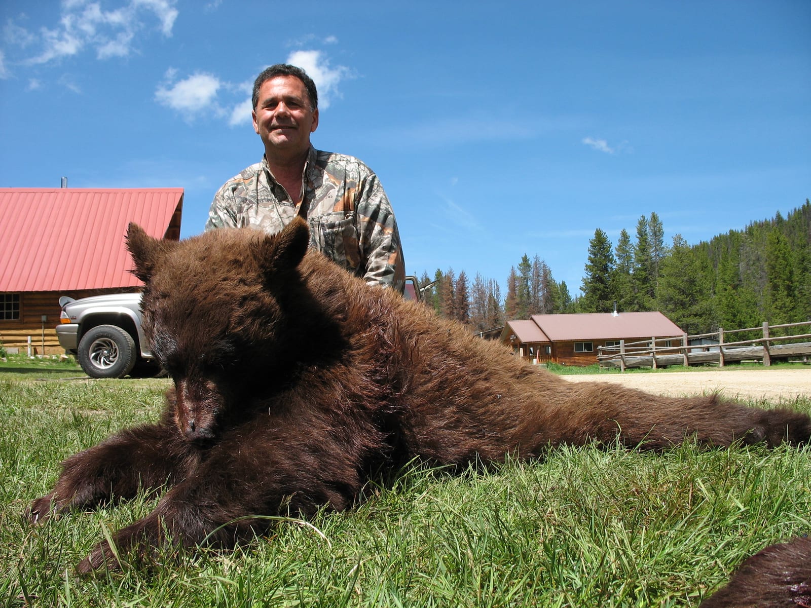 A hunter poses with a harvested black bear on a grassy field with buildings and trees in the background.