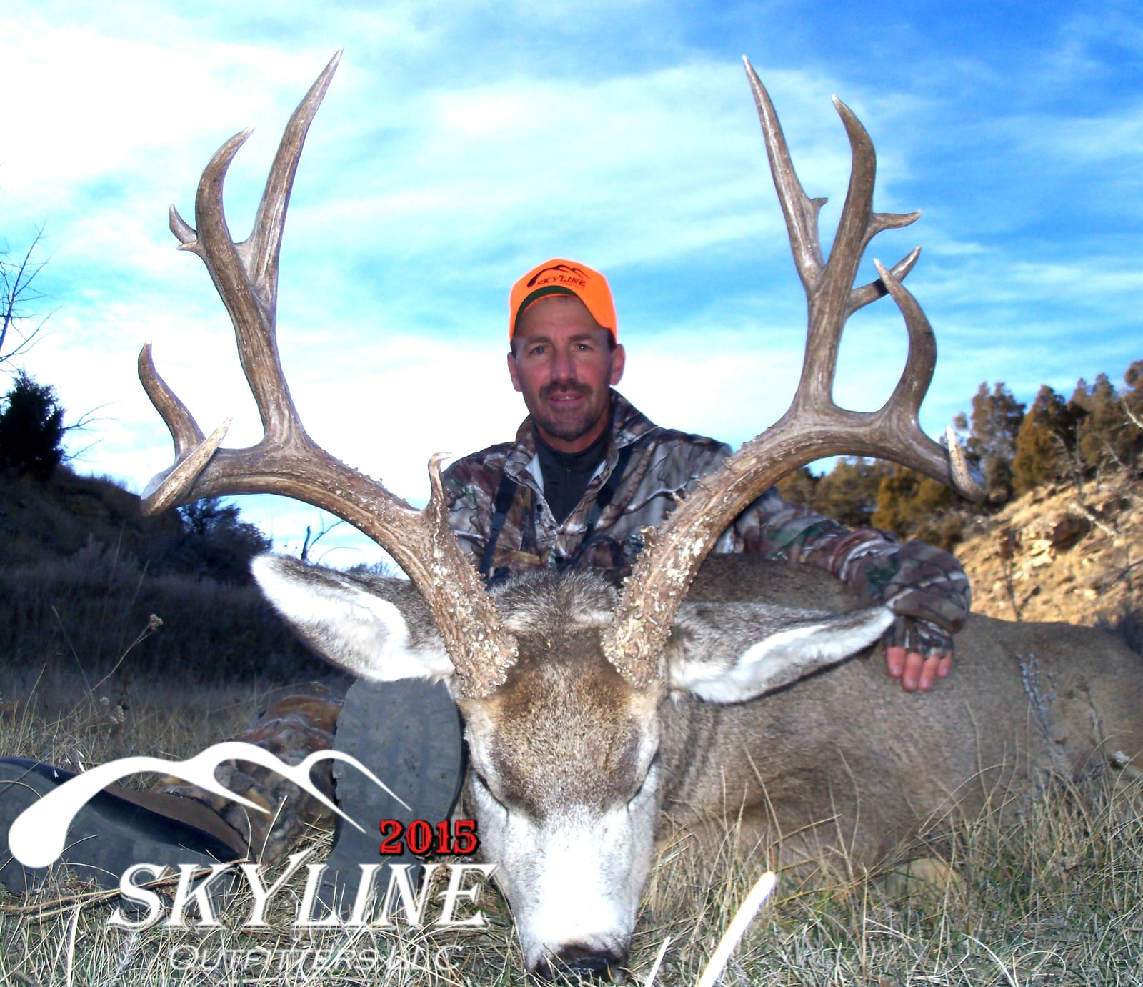 A hunter in camouflage and an orange hat poses with a large harvested mule deer.
