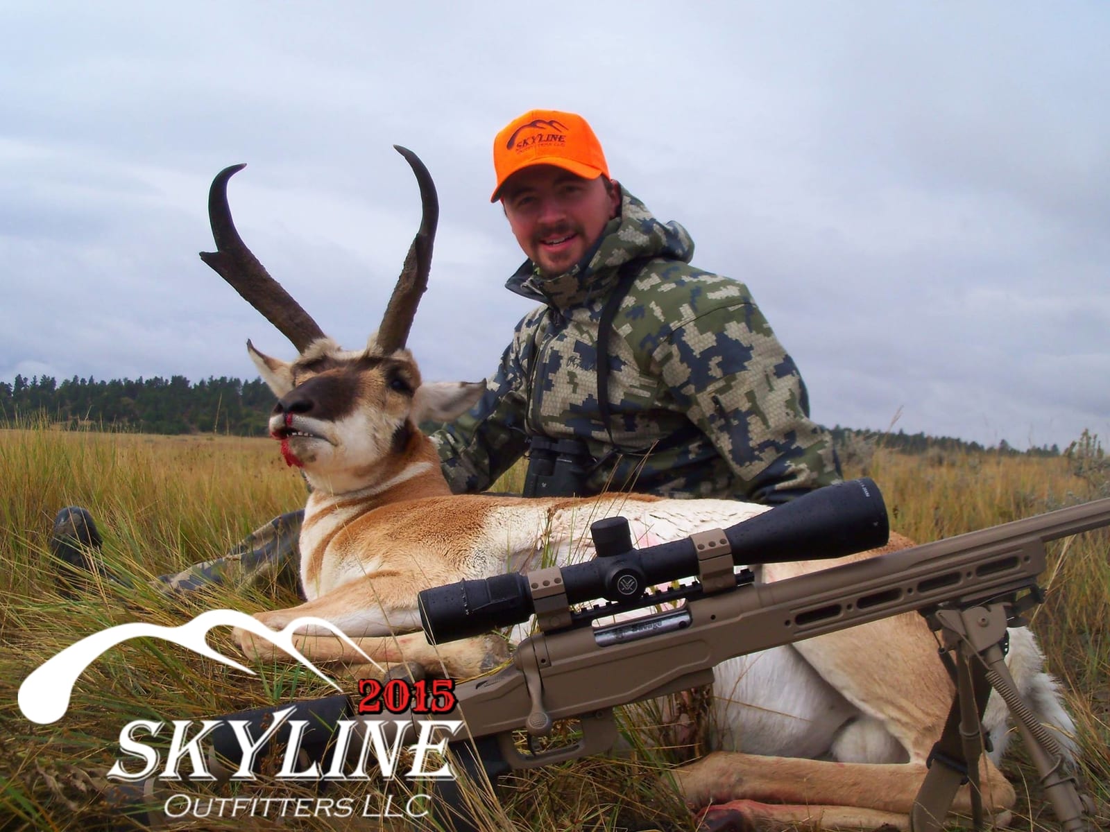 A hunter poses with a harvested pronghorn and a rifle in a grassy field.