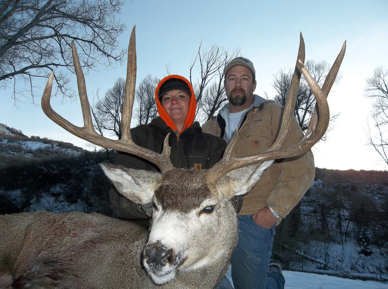 Two hunters pose with a harvested mule deer in a snowy, treed landscape.