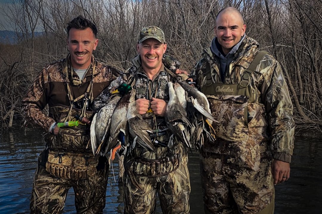 Three hunters in camouflage pose with their harvested ducks, including mallards, green-winged teal, and northern pintails, in a marshy area.