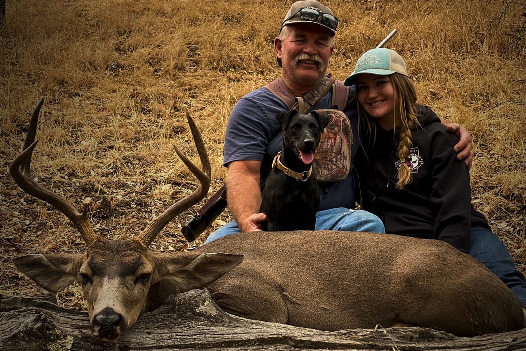A man, a girl, and a dog pose with a harvested mule deer.