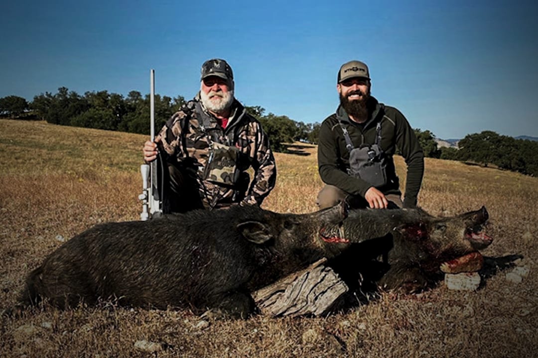 Two hunters pose with a harvested wild boar in a field.