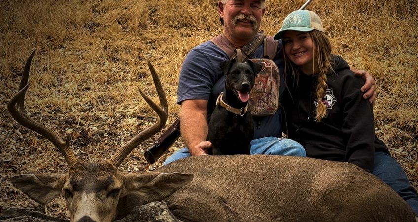 A man, a woman, and a dog pose with a harvested mule deer.