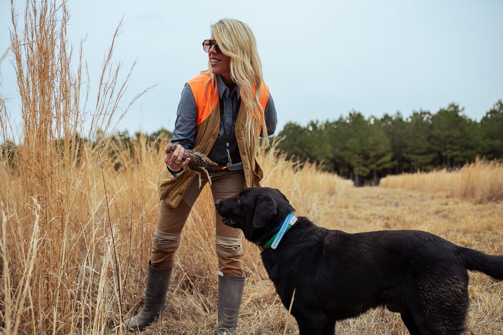 A woman in hunting attire holds a harvested ring-necked pheasant while a black dog stands beside her in a field.