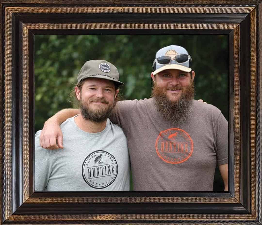 Two men, one with a beard, smile and pose for a photo, with one man's arm around the other.