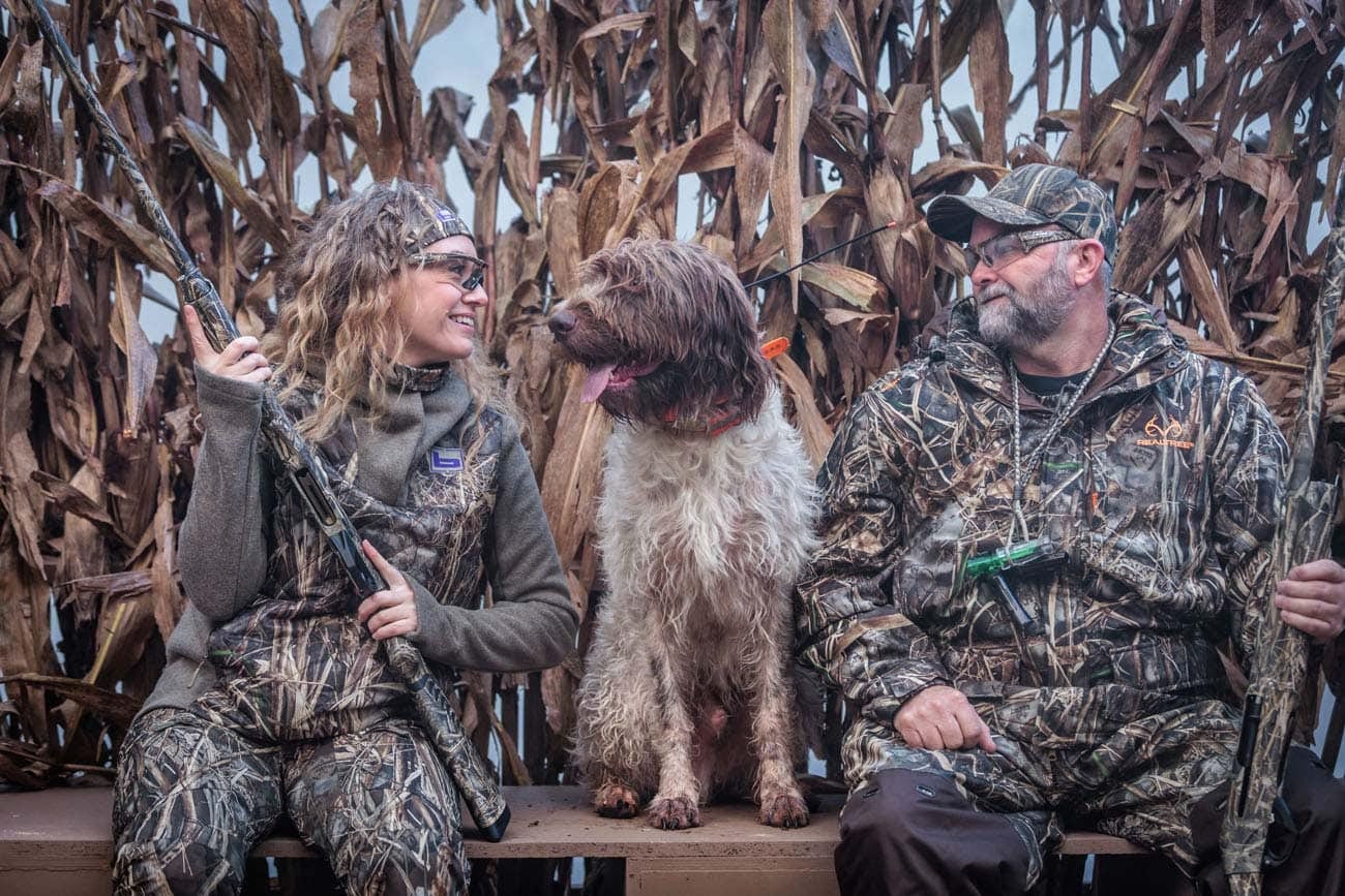Two hunters in camouflage sit with a hunting dog in a blind made of dried corn stalks.