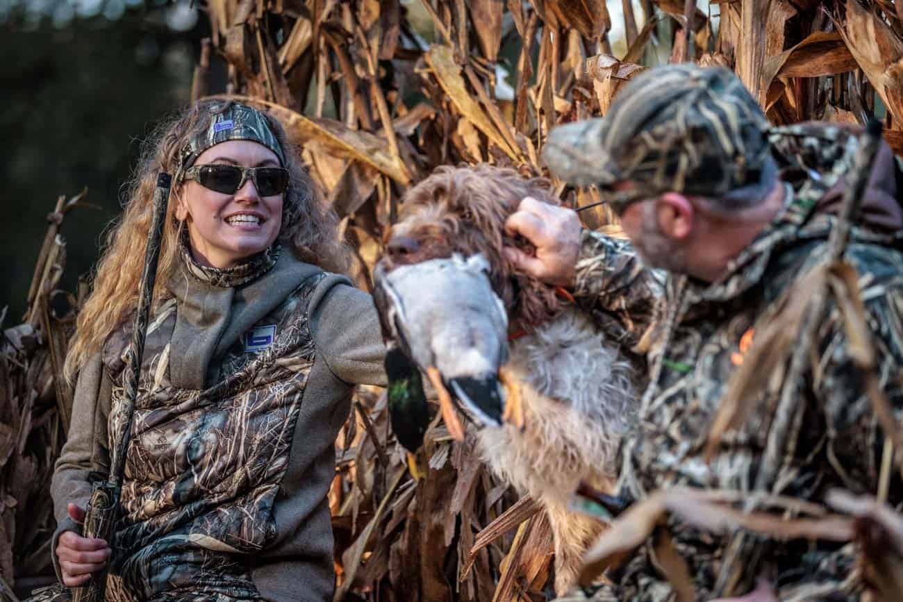 A woman and a man in camouflage with a hunting dog and a harvested mallard duck in a field.