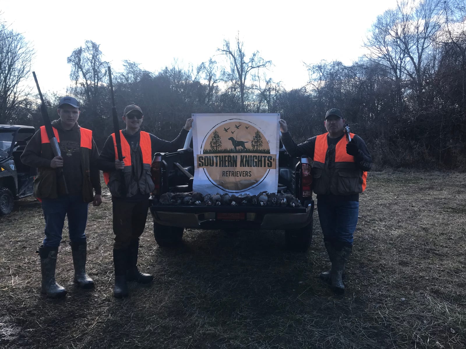 Three hunters pose with shotguns and a banner, standing next to a truck bed filled with harvested birds.