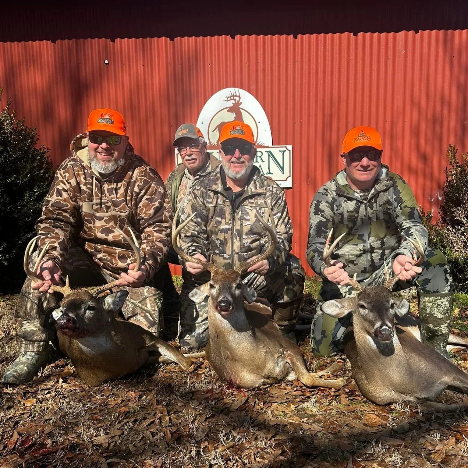 Four men in camouflage hunting gear posing with three deer they have hunted, holding antlers, in front of a red barn with a logo that features a deer head.
