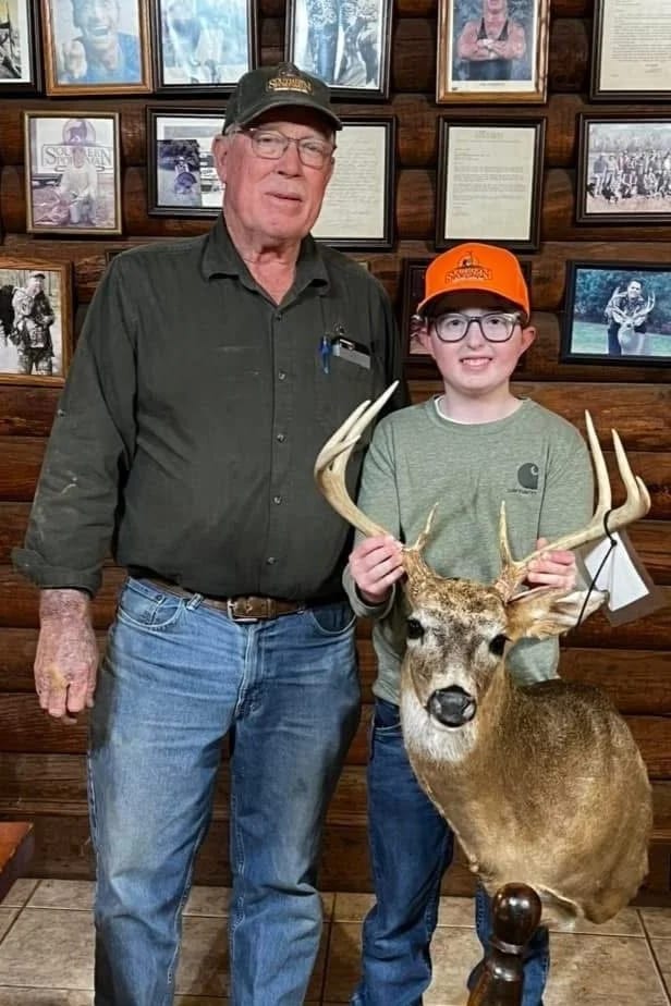 An elderly man and a young boy posing with a large deer head with antlers in an indoor setting with framed photos and documents on the wooden wall behind them.