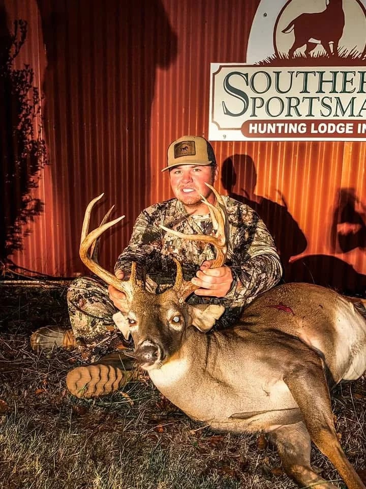 A man in camouflage clothing holding a large deer with antlers, posing in front of a red metal wall with a sign that reads 'Southern Sportsman Hunting Lodge'.