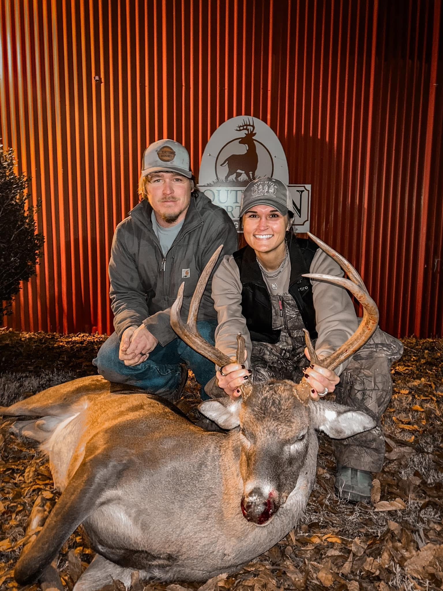 A man and woman kneeling next to a freshly hunted deer with large antlers, smiling at the camera, with a wooden wall and a sign with a deer logo in the background.