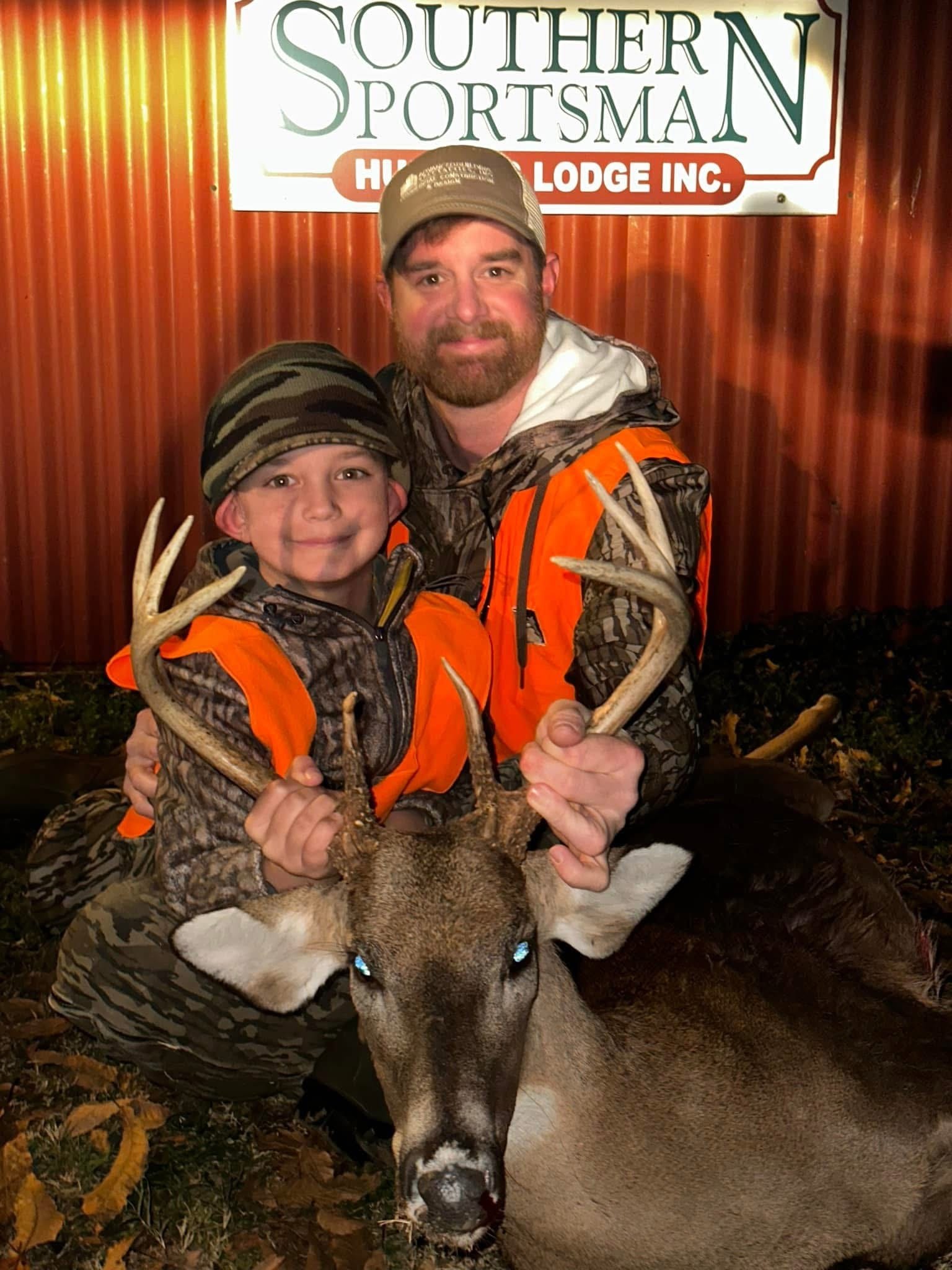 A man and a young boy in camouflage hunting gear and orange vests, holding a deer with antlers, posing in front of a red metal building with a sign that reads 'Southern Sportsman Hunting Lodge Inc.'
